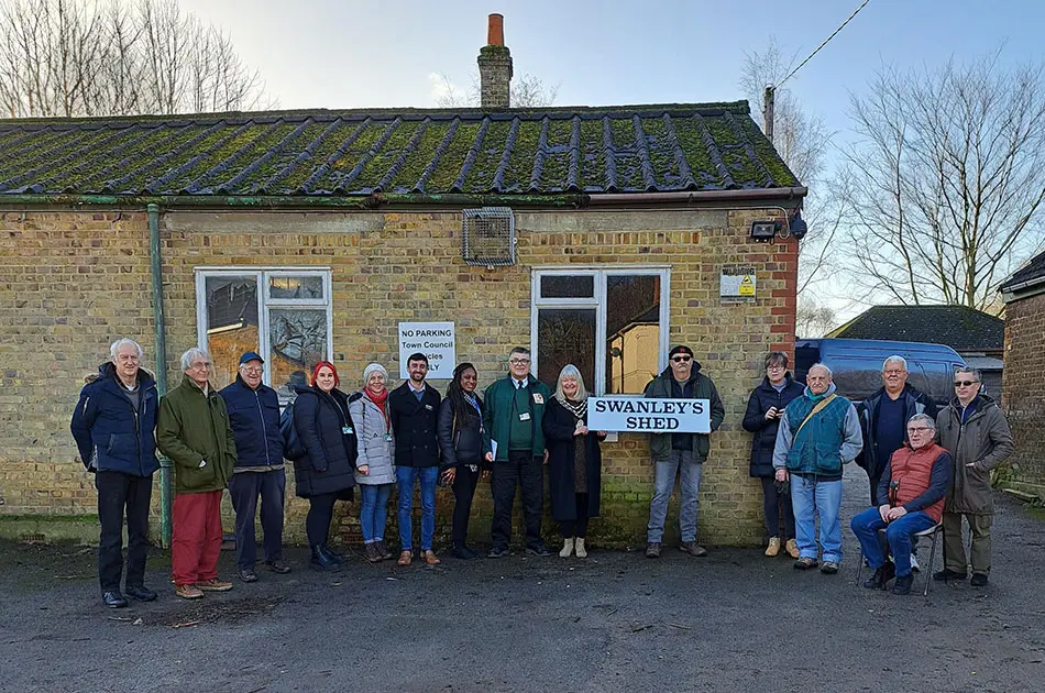 A group of people stand smiling outside a small brick building with a mossy roof. Two people hold a Swanleys Shed sign, celebrating one of the latest Shed Launches. Trees and a clear sky are in the background.