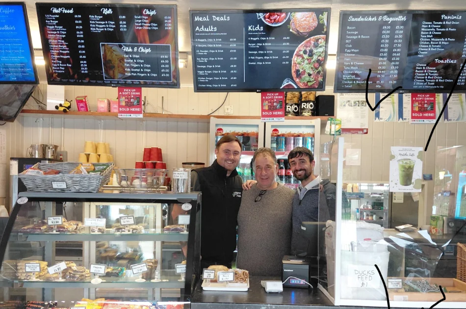 Three owners smile behind the counter of Park Café, with digital menu boards displaying food items and prices overhead.
