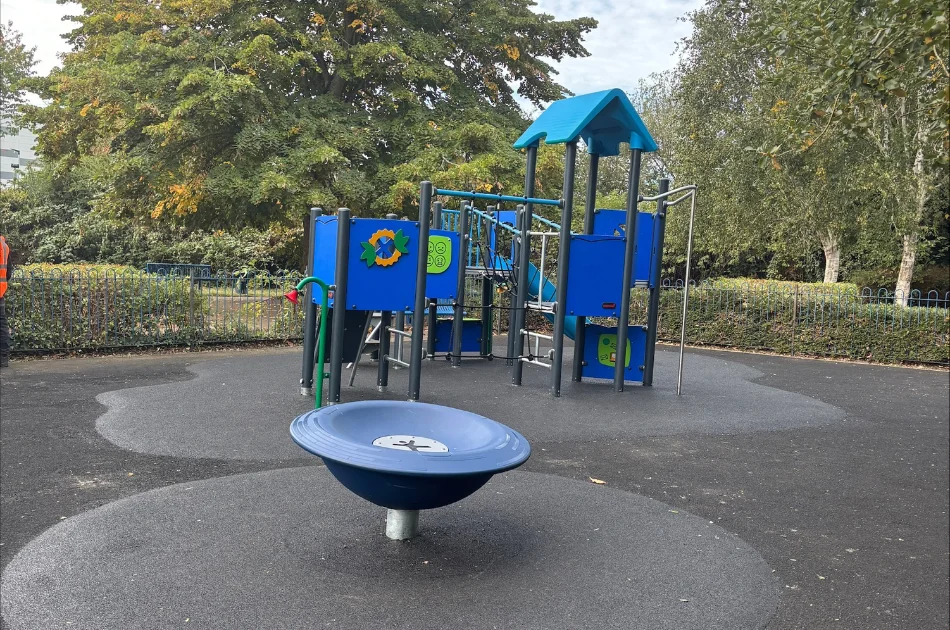 A playground in the Hilda May Playground featuring a blue multi-level play structure with a slide and a spinning bowl seat in the foreground.